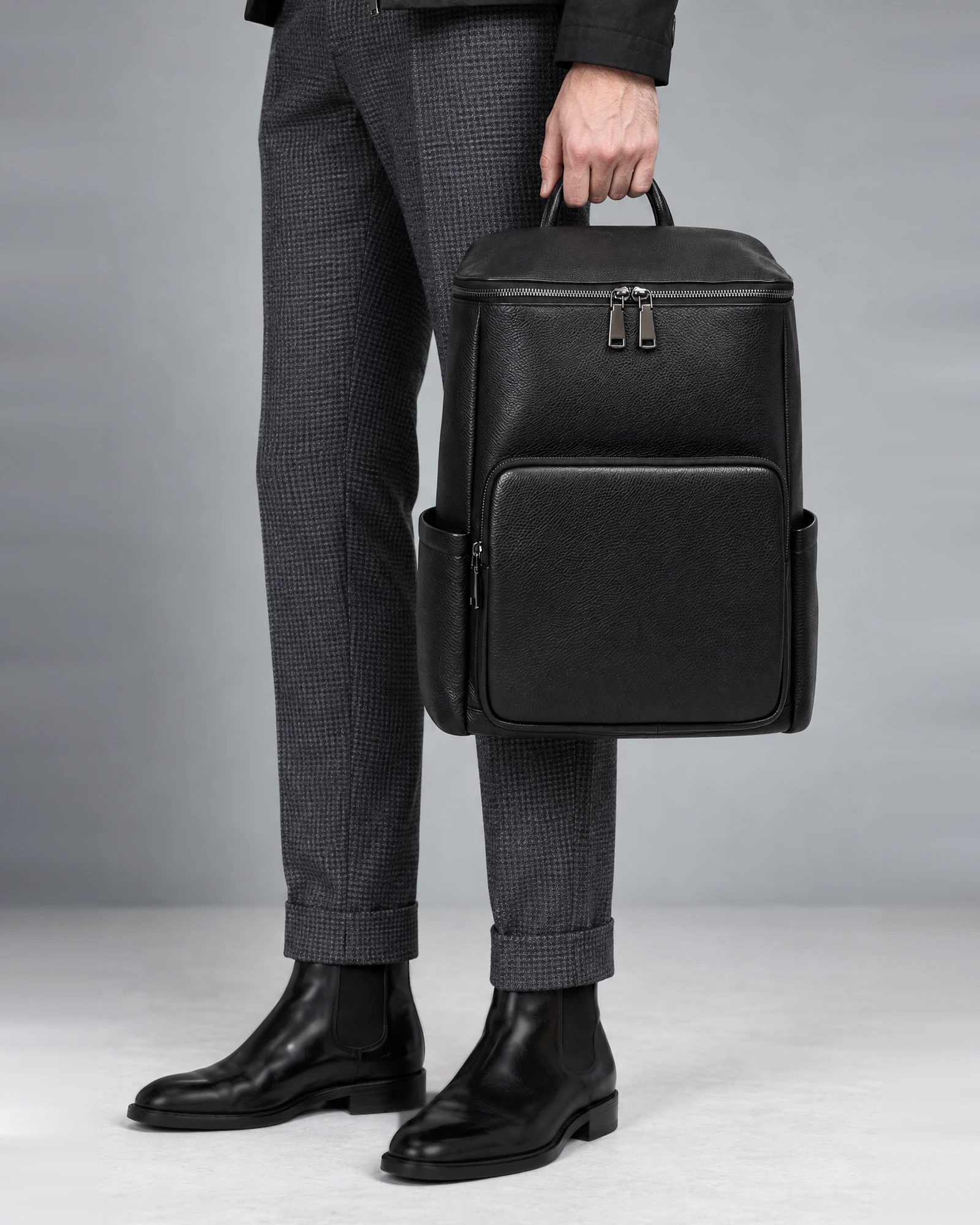 Le sac à dos LC/01 tenu à la main par sa poignée, homme en veste noire et pantalon gris, vue de trois-quarts sur fond gris